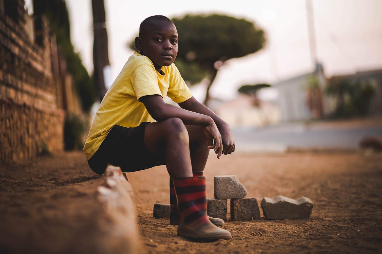 our-story A young boy in a yellow shirt sits outdoors on a quiet street during summer.
