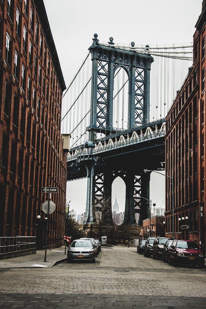 our-mission View of Manhattan Bridge between red brick buildings, with cars parked on the street.